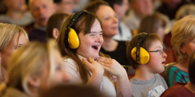 A girl sits in a theatre auditorium surrounded by other children and adults. She is wearing a pair of yellow ear defenders and has her hands raised to her chin in excitement.