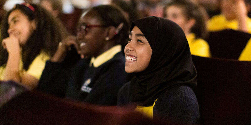 A schoolgirl is seated in a theatre surrounded by classmates, all wearing black and yellow uniforms. The girl wears a hijab and is smiling broadly.