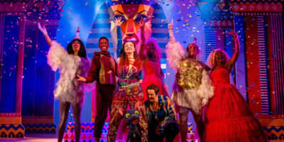 A diverse cast of 7 performers stand on stage with hands in the air in joyous celebration. The performer at the front of the group is operating a large puppet of a cat, and the set and costumes are in a fantasy Ancient Egyptian theme. Confetti falls from above onto the performers.