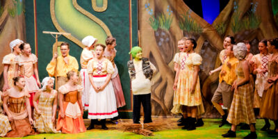 Children perform in Shrek The Musical. One child in the centre is dressed in a green hat with green facepaint, and two groups of children either side wear medieval-style costumes and are singing.