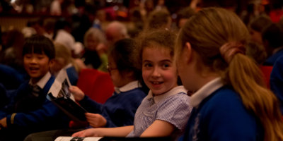 Image of two young pupils sat in theatre seats talking to each other while surrounded by other students at Swan Lake Schools' Matinee © 2022 in the Royal Opera House. Photographed by Rachel Cherry