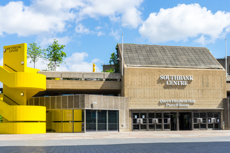Image is of the Queen Elizabeth Hall at the Southbank Centre. The building is 70s brutalist in design and there is a bright yellow concrete staircase on the left
