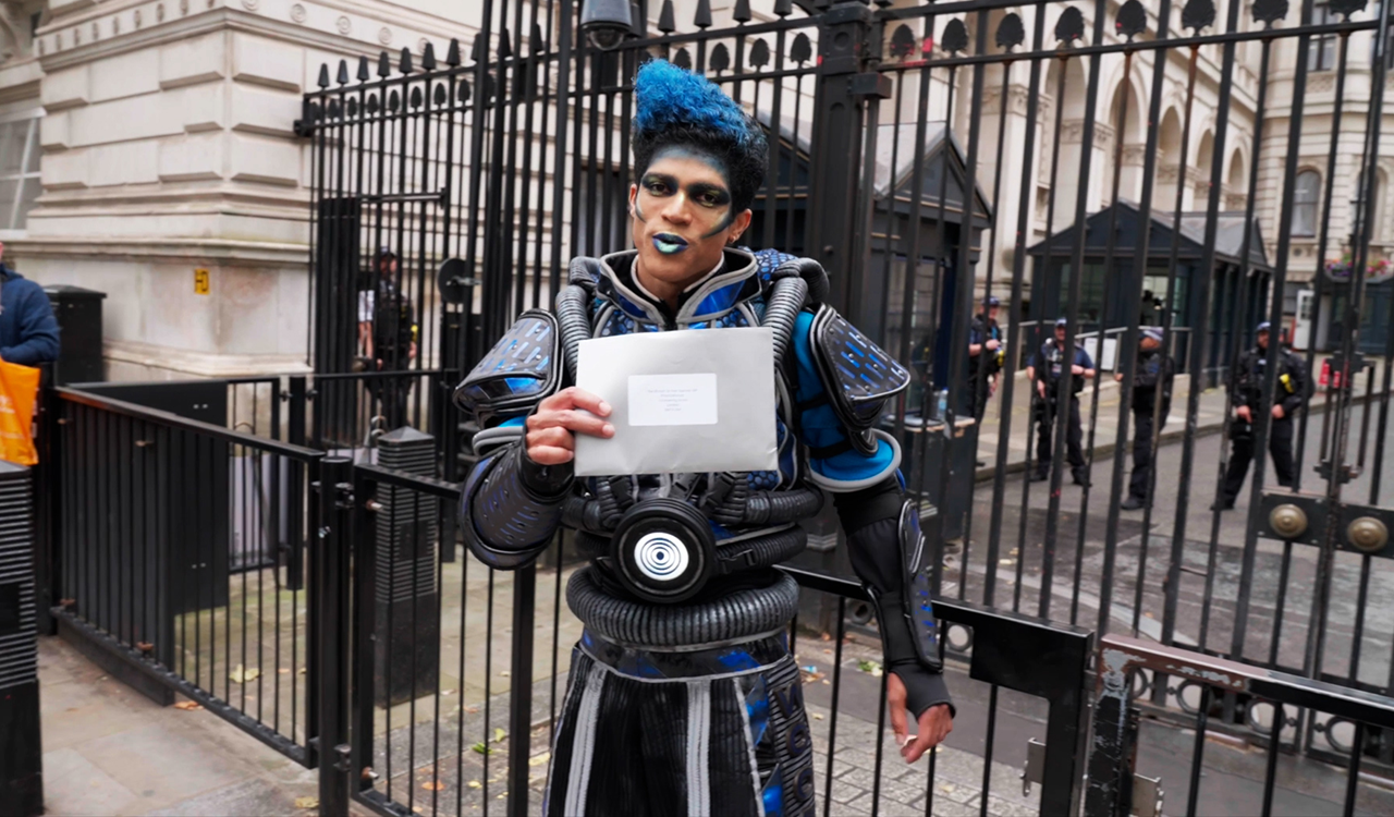 Cast member from Starlight Express with bright blue hair and make-up holds out a silver envelope addressed to 10 Downing Street