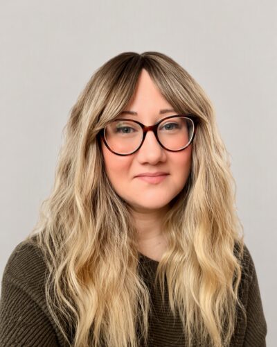 A headshot photo of Becci. She is a caucasion woman with blonde, wavy hair, black round glasses and is wearing a dark green jumper.