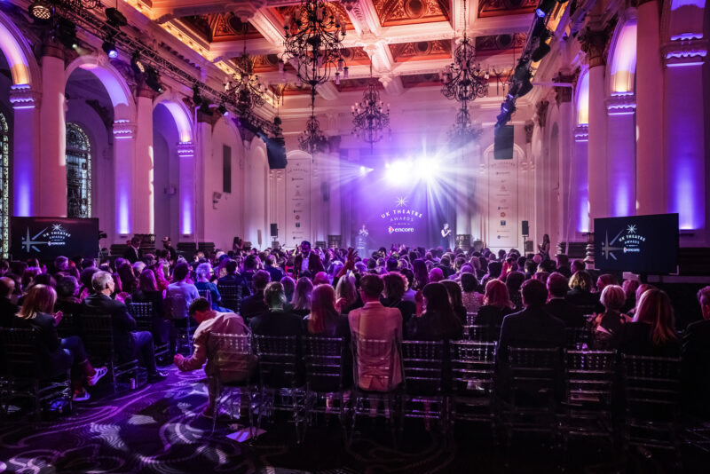 A wide angle of a room full of people all looking towards a stage. There is lights and smoke and the room appears to be pink and purple because of the light.