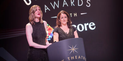 Two women are standing on stage behind a podium at an awards ceremony. The woman on the left (Claire Walker) is tall with blonde hair in waves, wearing a sleeveless, sparkly black dress. The woman on the right (Hannah Essex) has medium brown hair and is wearing a short-sleeved black dress with gold accents. Visible on both the podium and backdrop is the 'UK Theatre Awards with Encore' logo.