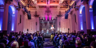 An audience is seated in an ornate hall with high ceilings, chandeliers and arches lit in purple and blue. On stage, Jenny Sealey is receiving an award, with a large screen behind her displaying her image and the text "Winner of Outstanding Contribution to British Theatre." She is speaking at a microphone in the centre of the stage and wearing a long green scarf. To Jenny's right, the host Faye Tozer stands behind a podium. To Jenny's left is a BSL interpreter dressed in black.