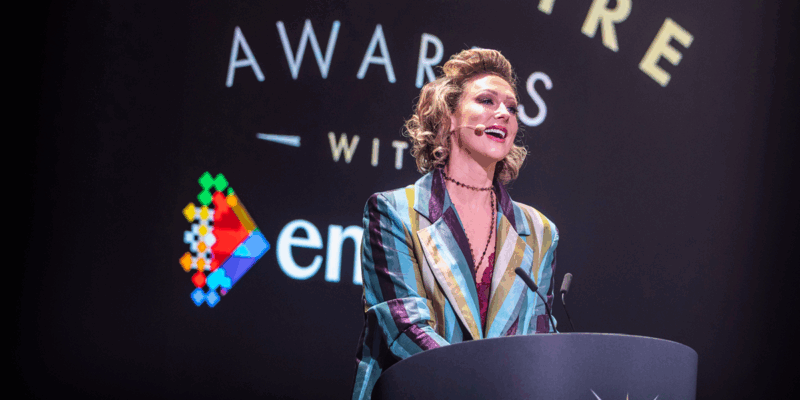 Faye Tozer speaks at a podium during an awards ceremony, wearing a striped jacket and headset microphone.