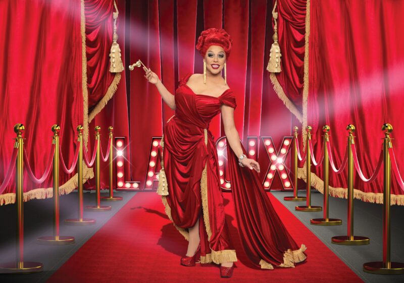 La Voix, wearing a red evening gown, stands on a red carpet in front of red theatre curtains, with gold velvet ropes and illuminated letters spelling “LA VOIX” behind her.